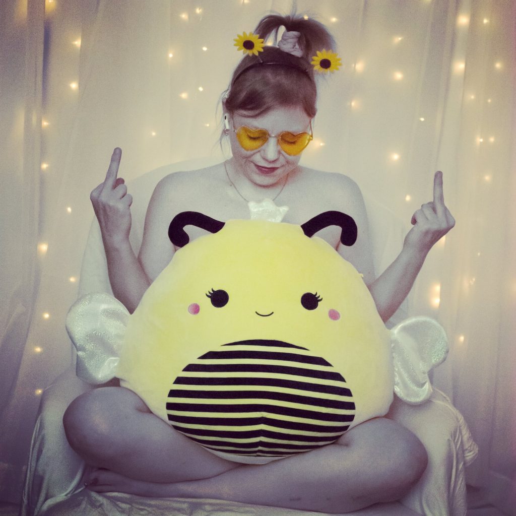 A photo of Sunny Crittenden sitting in a chair draped with white fabric, with a large yellow, stuffed bee in her lap, giving the finger with both hands. Sunny is nude except for her yellow heart-shaped sunglasses, yellow sunflower wheelie bopper headband, and airpods. Her eyes are closed.