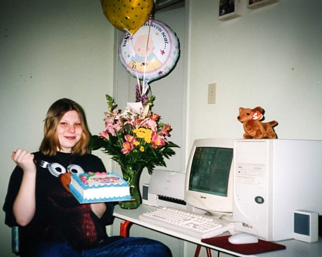 Photo of Sunny Crittenden in 1998 the day of her "cyber baby shower". She is sitting in front of an old clunky desktop computer, holding a cake with indecipherable writing on it in one hand and a fork with the other. She has shoulder length ash blonde hair and is wearing an oversized black t-shirt with the sesame street character Elmo on it. On the desk in front of her there is a vase with assorted flowers, and baby shower themed balloons. Sunny is in her 3rd trimester of pregnancy with her 1st child, Madison. There is a weiner dog beanie baby sitting on top of the computer tower, indicative of the times.