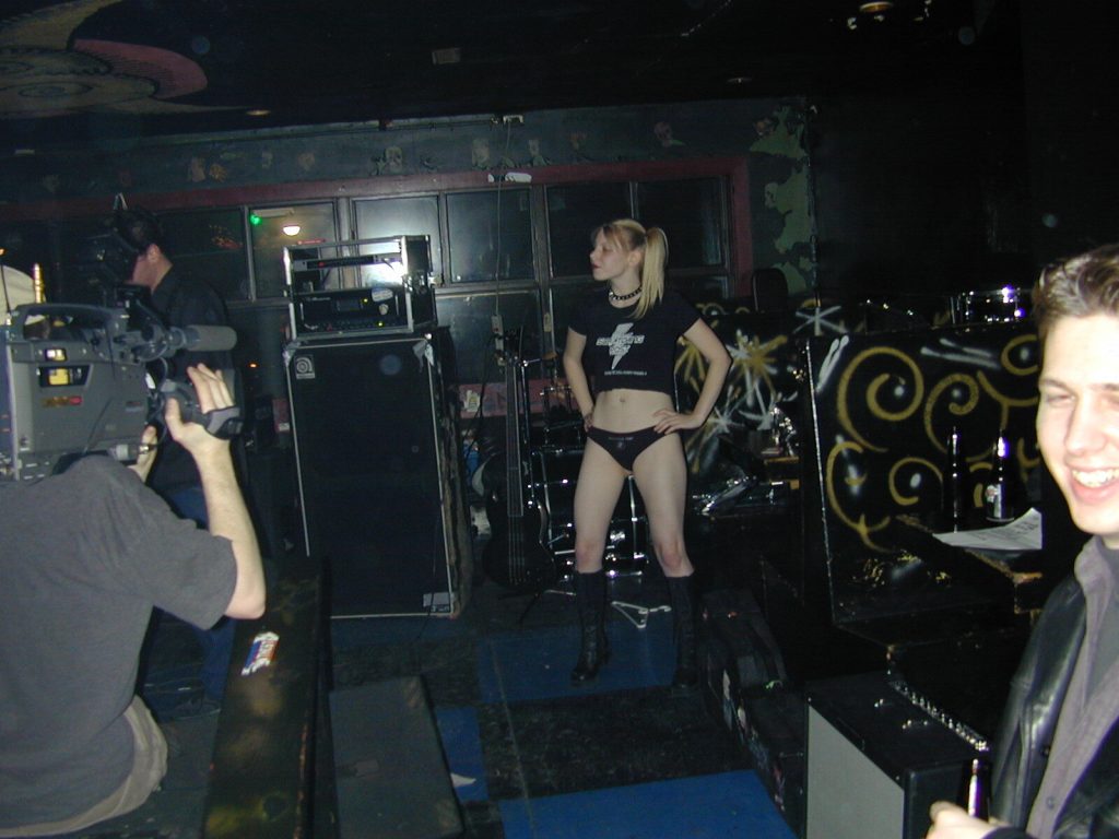 A photo of Sunny Crittenden on stage, wearing her Scratching Post Panty Girl uniform and being filmed by the CBC at Sneaky Dee's in Toronto circa fall 2001.