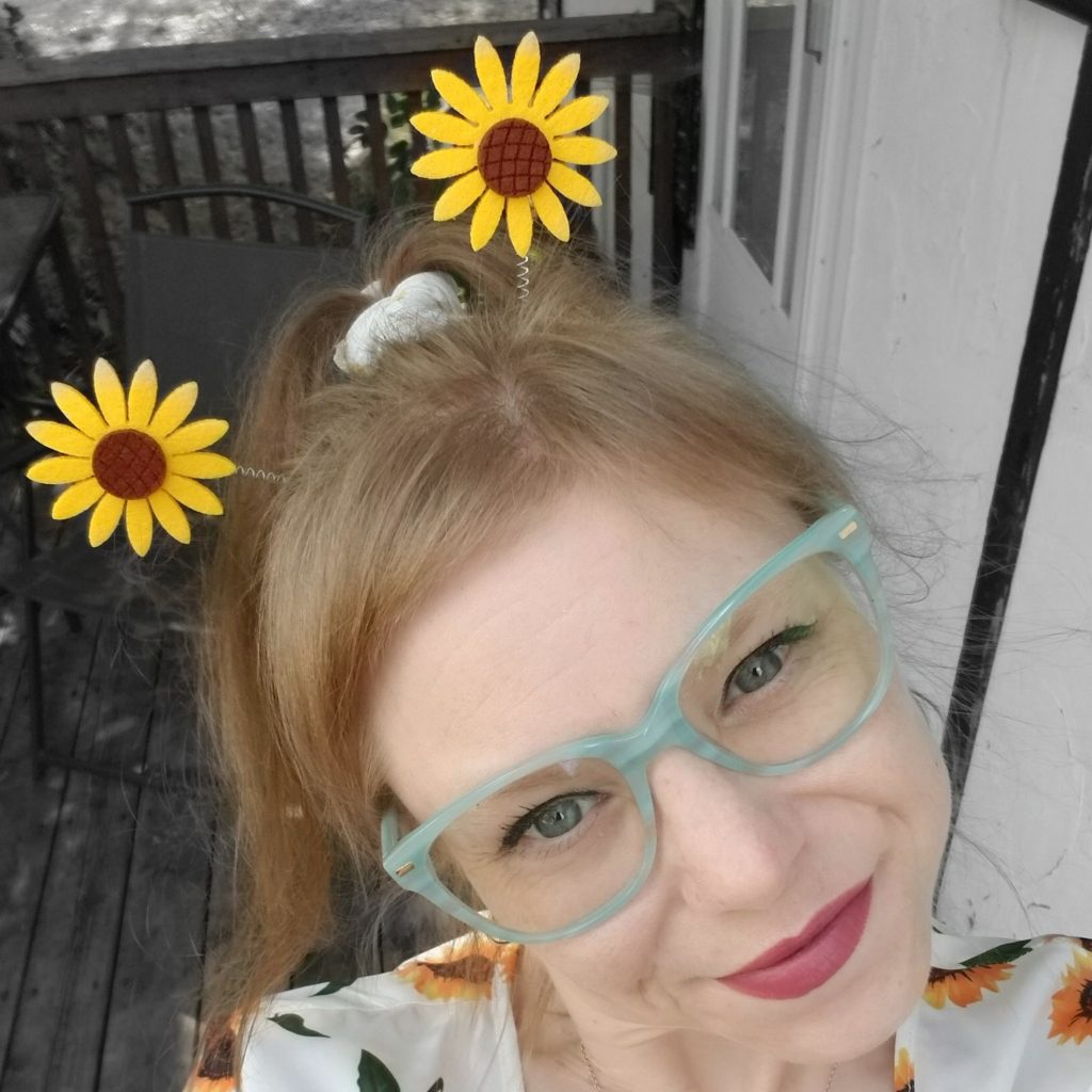 Selfie photo of Sunny Crittenden smiling, wearing lipstick, her trademark baby blue glasses, a sunflower wheelie bopper headband, and a white dress with sunflowers on it. Her hair is tied up in a high ponytail with a matching sunflower scrunchie.