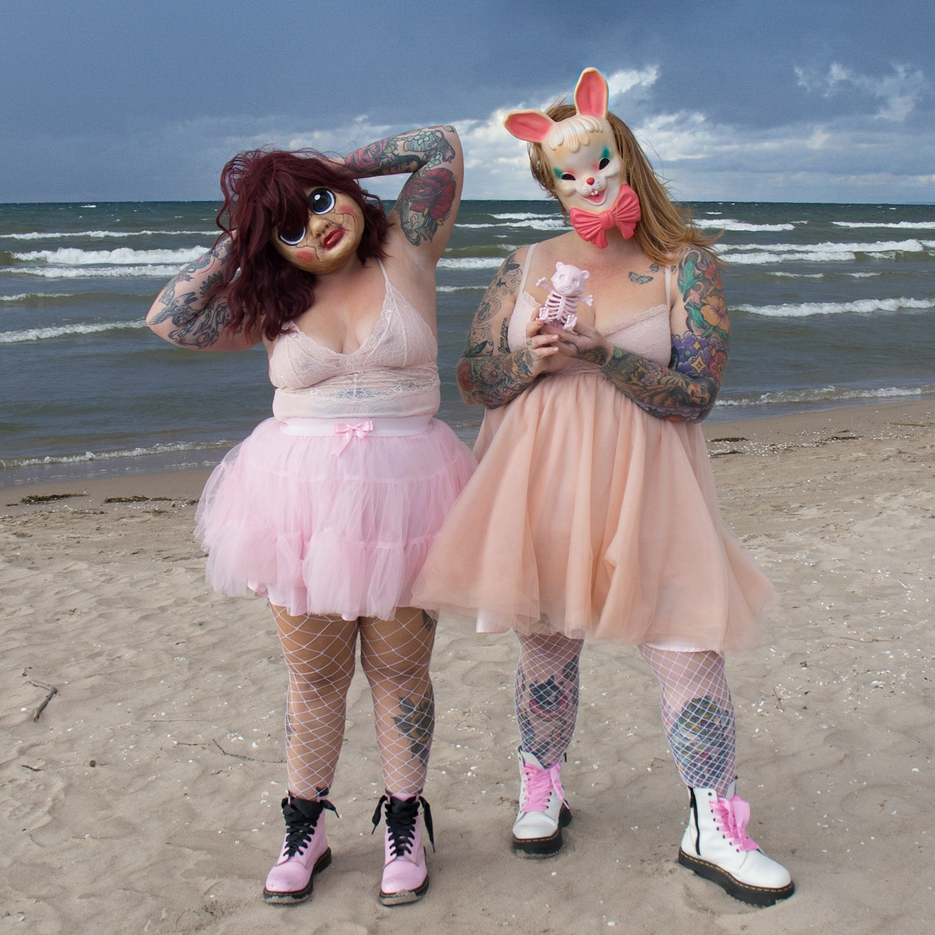 A colour photograph of two women wearing pink babydoll lingerie, pink and white Doc Martens, and creepy masks, standing on a moody beach, with their backs to the water. The sky is dark grey and stormy, and the waves are white and choppy. The water, being Wasaga Beach, is a green-ish blue. The woman on the right is holding a small, baby pink, plastic skeleton bear in both hands. This photo was chosen by Jones Soda to be on a Blue Bubblegum label on bottles to be sold everywhere Jones is sold.